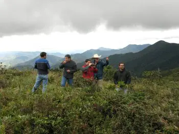 Group of people standing on a grassy hillside with mountains and a cloudy sky in the background, chatting and smiling.