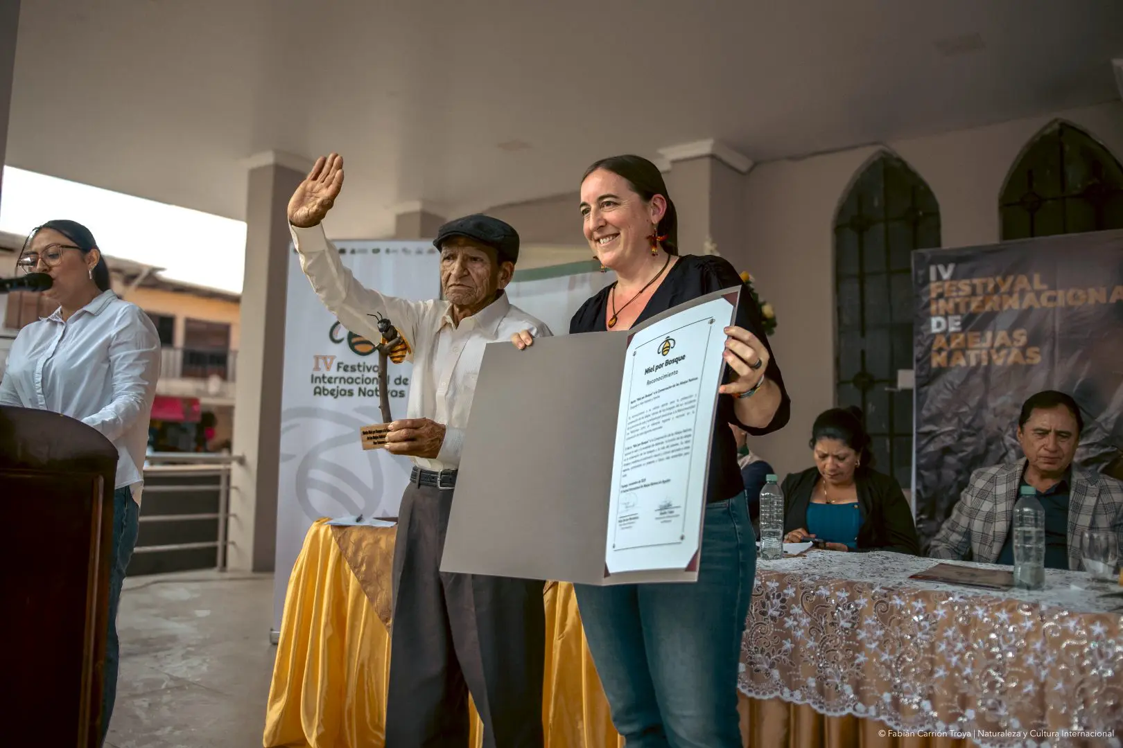 Man in white shirt and cap waves while receiving a framed certificate from a smiling woman at a ceremony on stage, bee festival banners in background