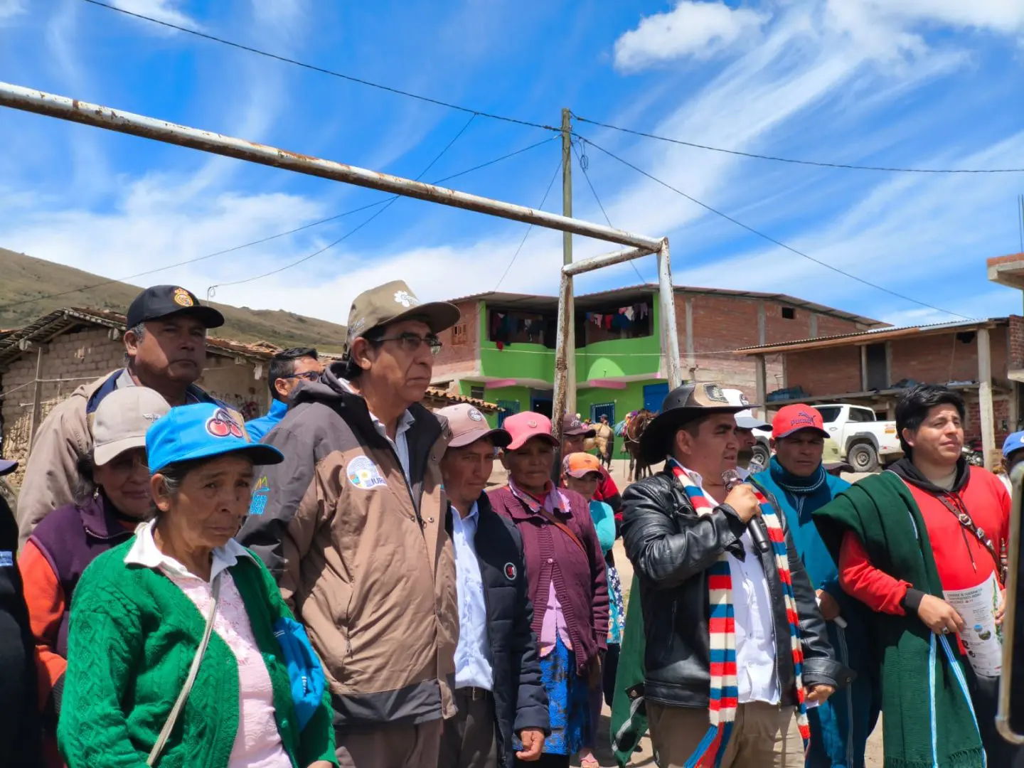 Group of villagers gathered outdoors in a rural town, some wearing hats and jackets, against a bright blue sky and building facades behind them.