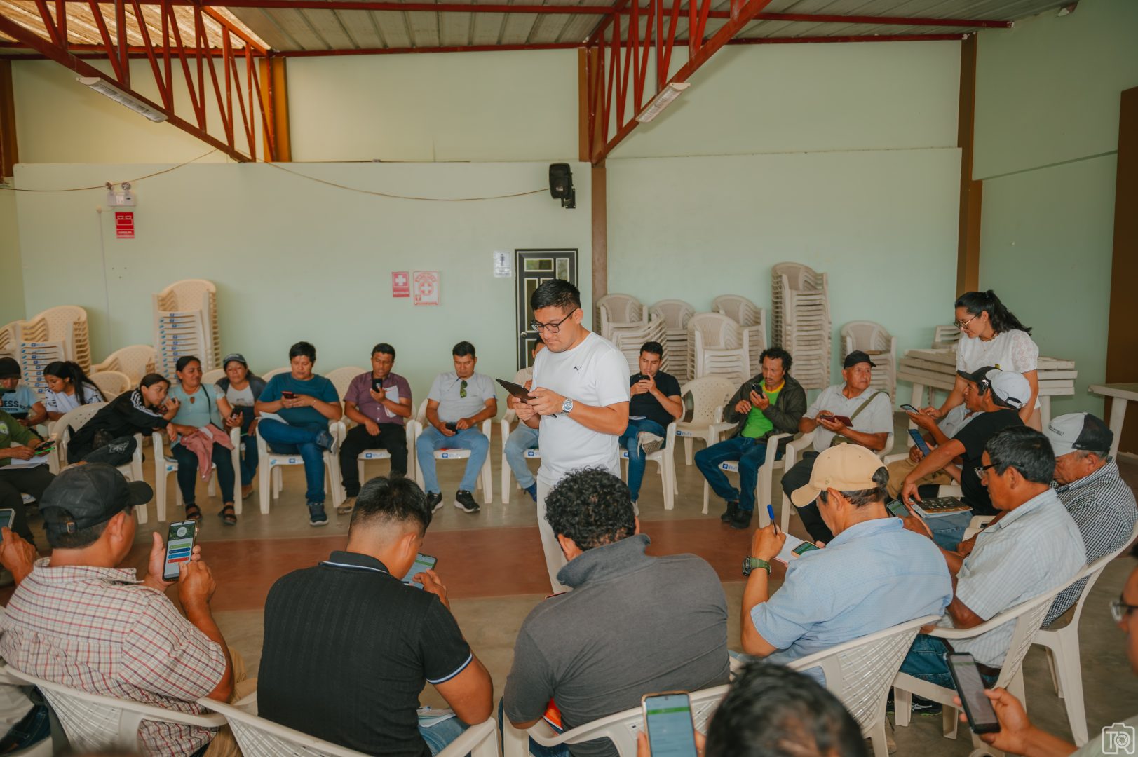 Group of people seated in a large circle inside a hall, many checking smartphones while a man stands in the center reading from a card or note believably leading the session.
