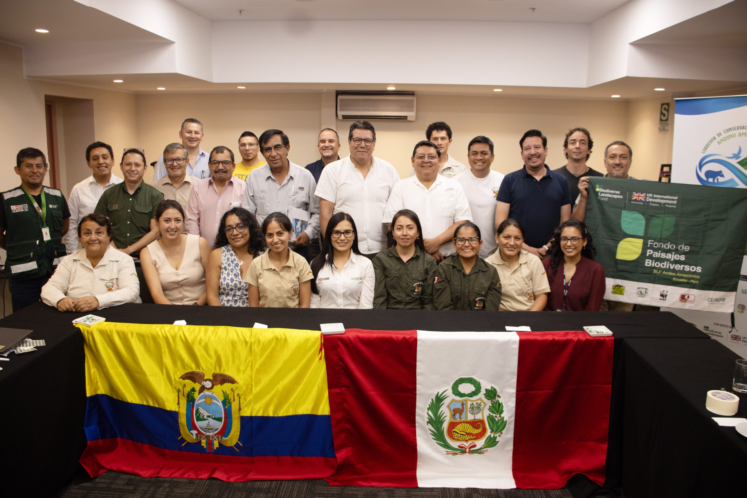 Group of diverse professionals posing for a formal photo at a biodiversity event, seated behind flags of Ecuador and Peru with banners in the background.