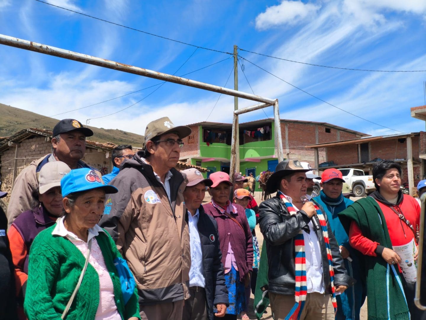 Group of villagers gathered outdoors in a rural town, some wearing hats and jackets, against a bright blue sky and building facades behind them.