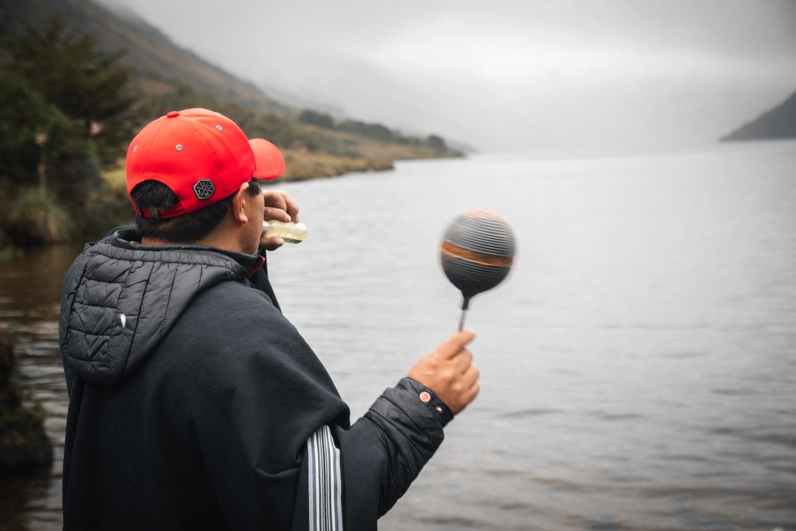 Man wearing a red cap and dark jacket stands at the edge of a lake, tossing a striped fishing float into the water on a cloudy day.