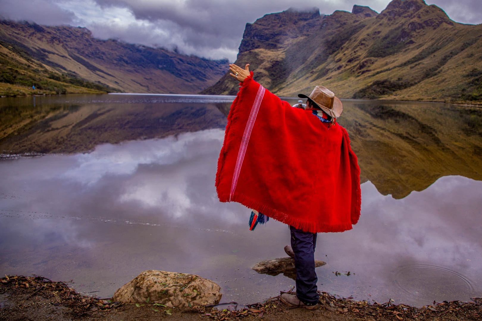 Person in a bright red poncho stands at a lake, raising one hand toward the misty mountains.